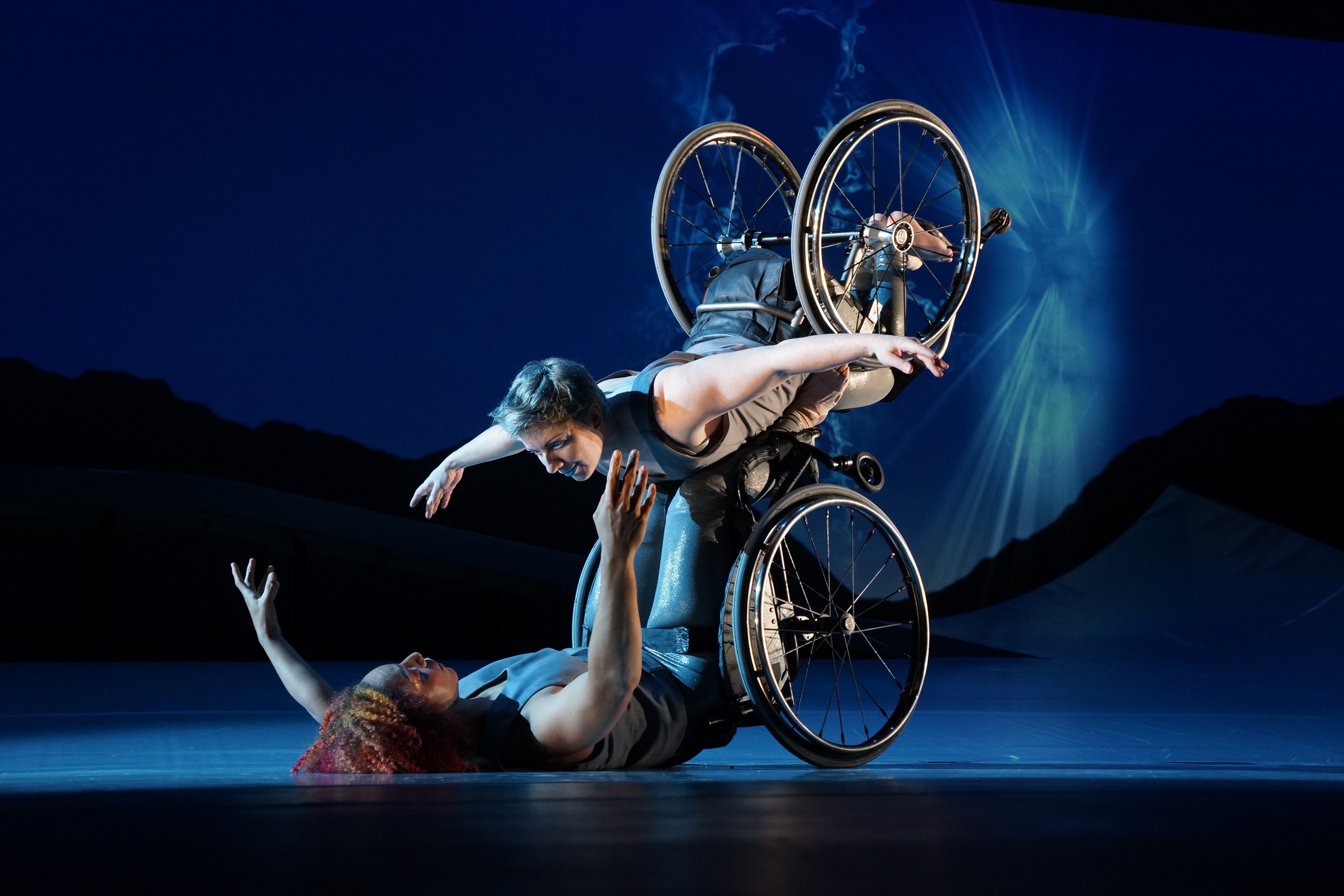 Laurel Lawson, a white woman with short cropped teal hair, is flying in the air with arms spread wide, wheels spinning, and supported by Alice Sheppard. Alice, a multiracial Black woman with coffee-colored hair, is lifting from the ground below. They are making eye contact and smiling. A burst of white light appears in a dark blue sky. Photo by Jay Newman / BRITT Festival.